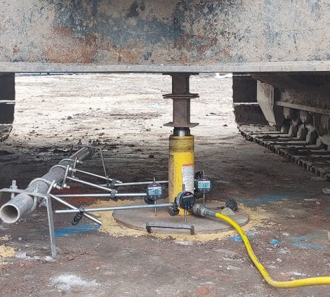 Plate load test setup beneath tracked excavator showing hydraulic jack, gauges and reaction beam